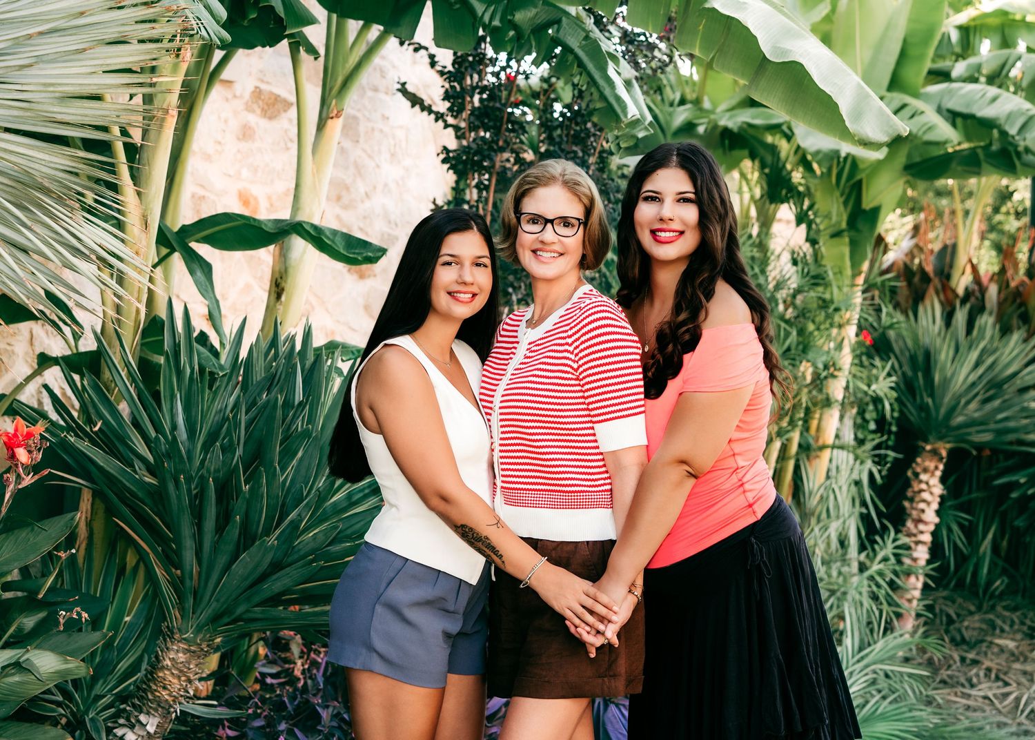 Three women smiling together outdoors surrounded by lush tropical greenery and palm plants in a bright, colorful setting.