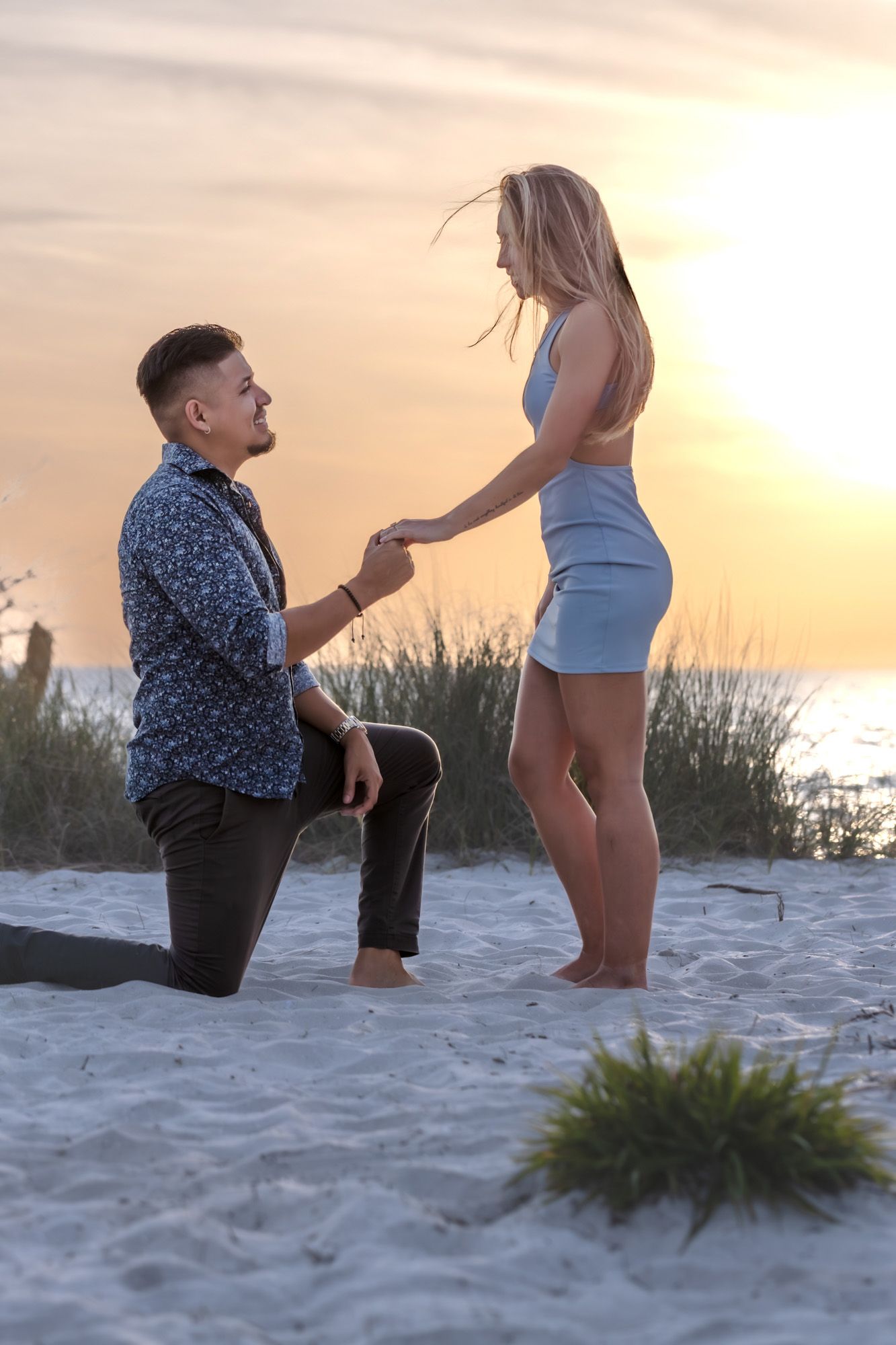 Man proposing on one knee to woman on sandy beach at golden sunset.