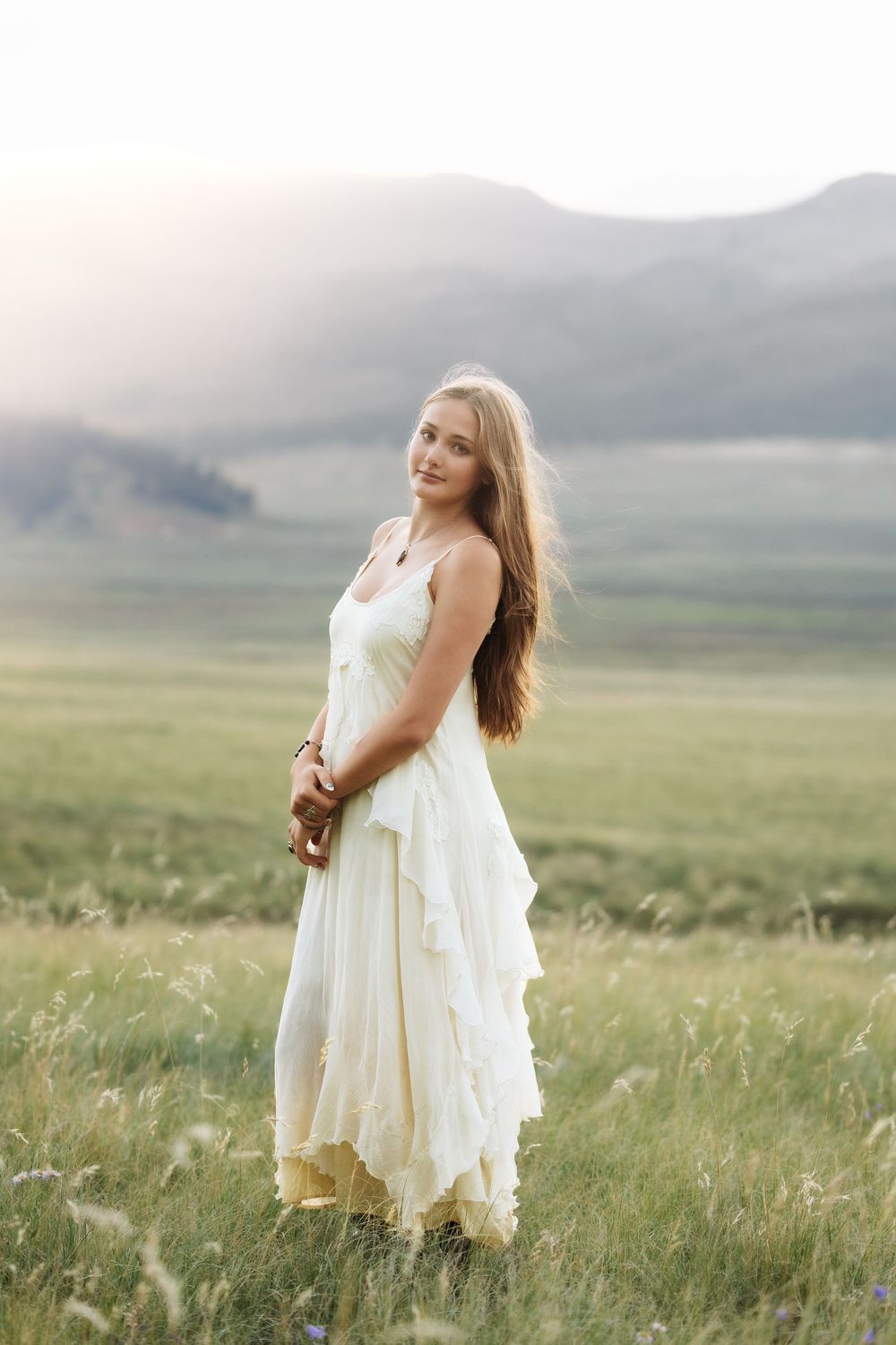 Young woman in white ruffled dress standing in green meadow with misty mountains in background.