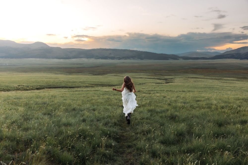 A woman in a white dress runs through a vast green meadow at sunset with mountains in the background.