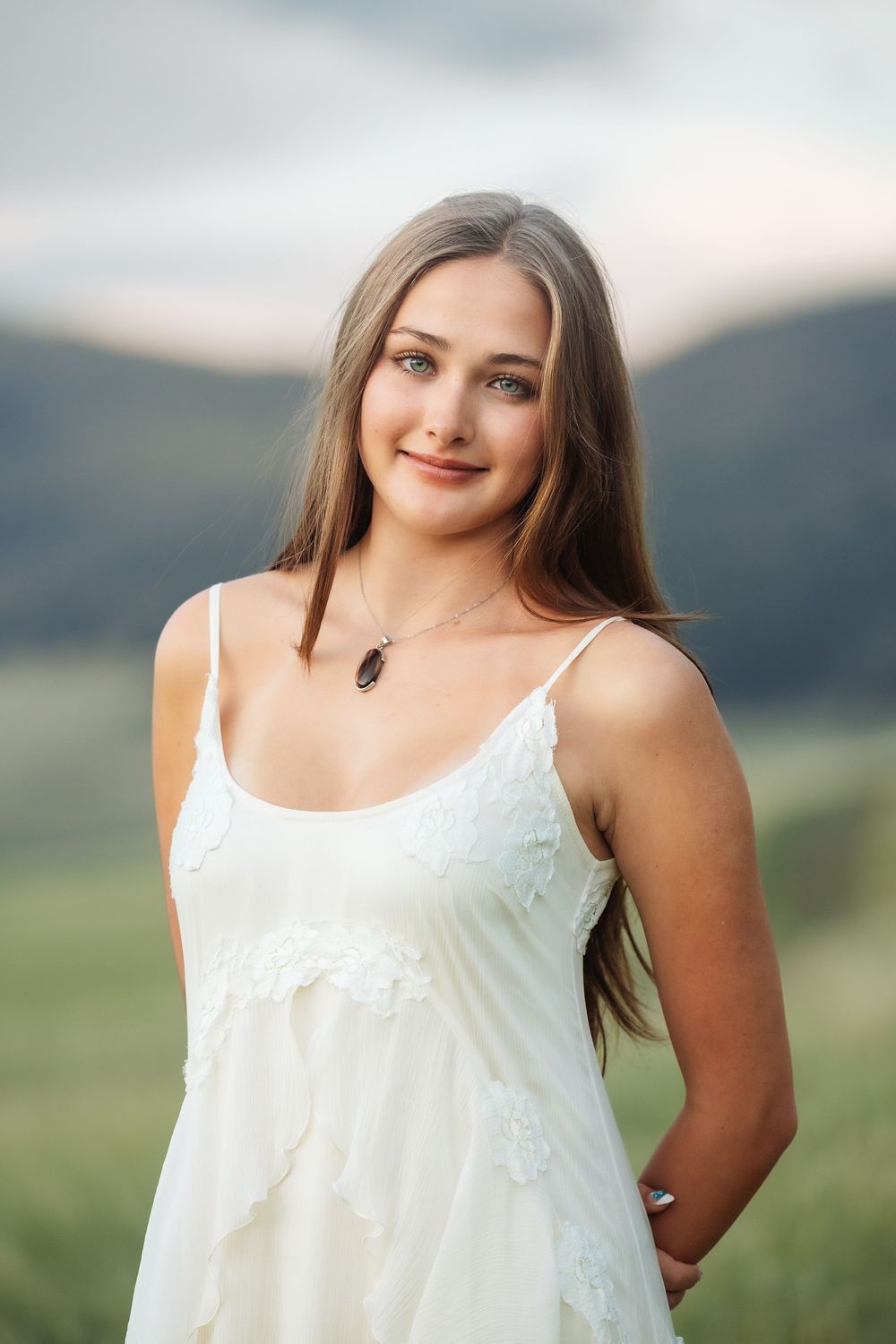 Young woman in white sundress with necklace smiling outdoors with mountain landscape backdrop.