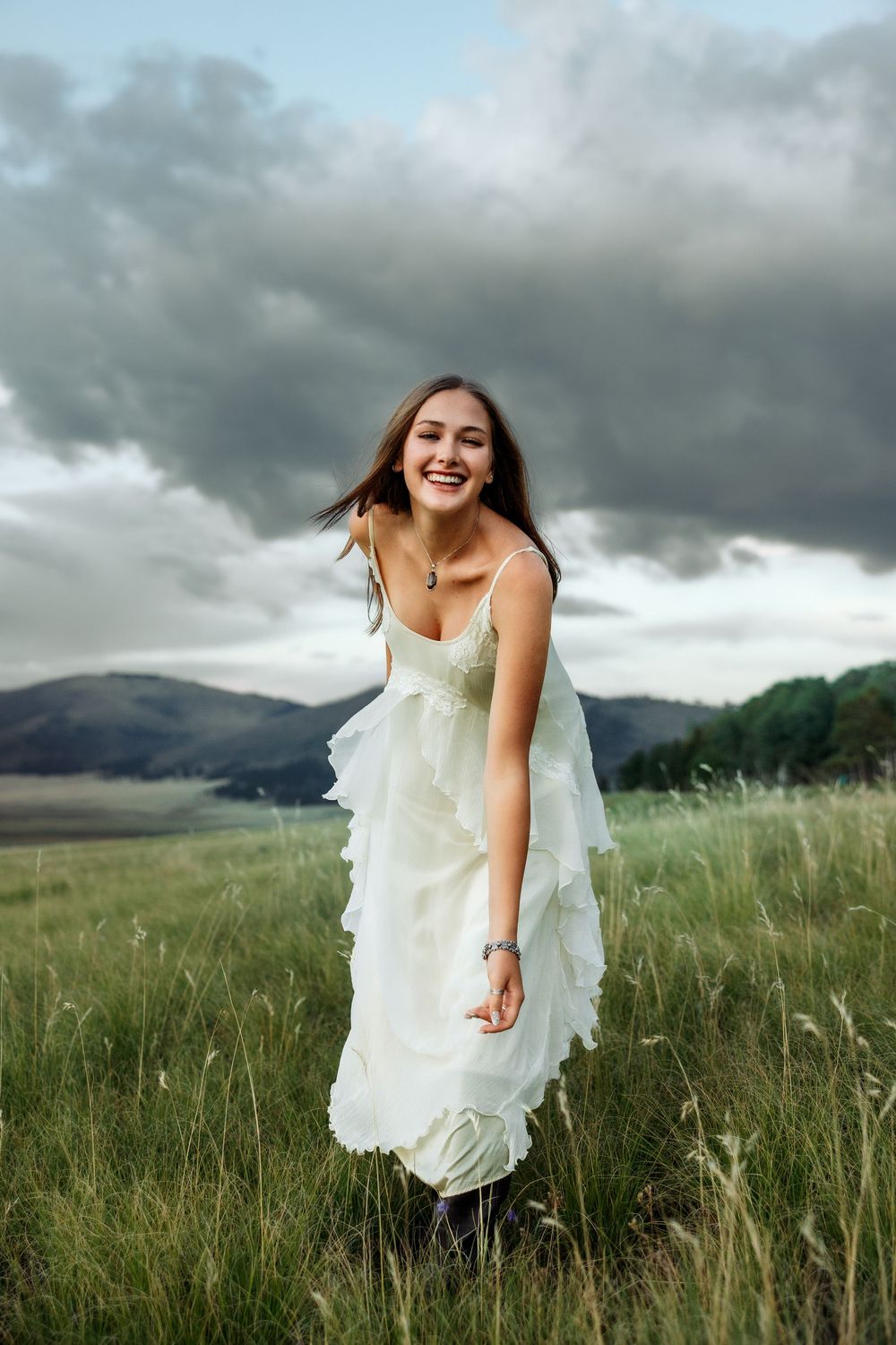 Young woman in white ruffled dress laughing in green meadow with mountains and dramatic cloudy sky.