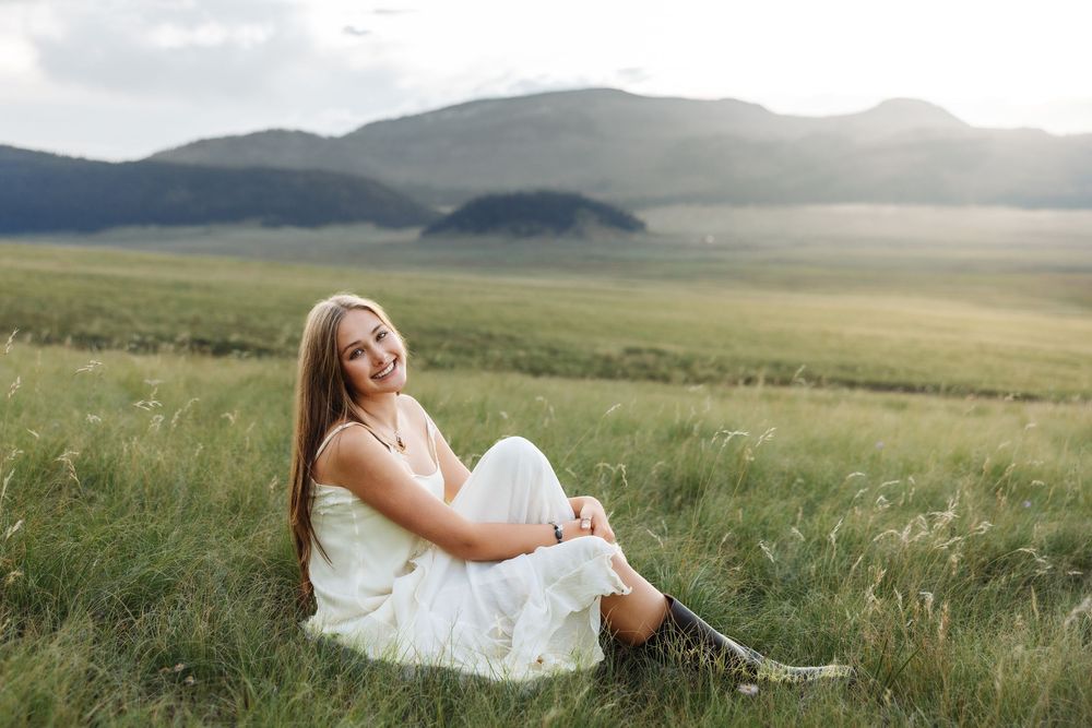 Young woman in white dress sitting in green meadow, smiling with mountain landscape in background.