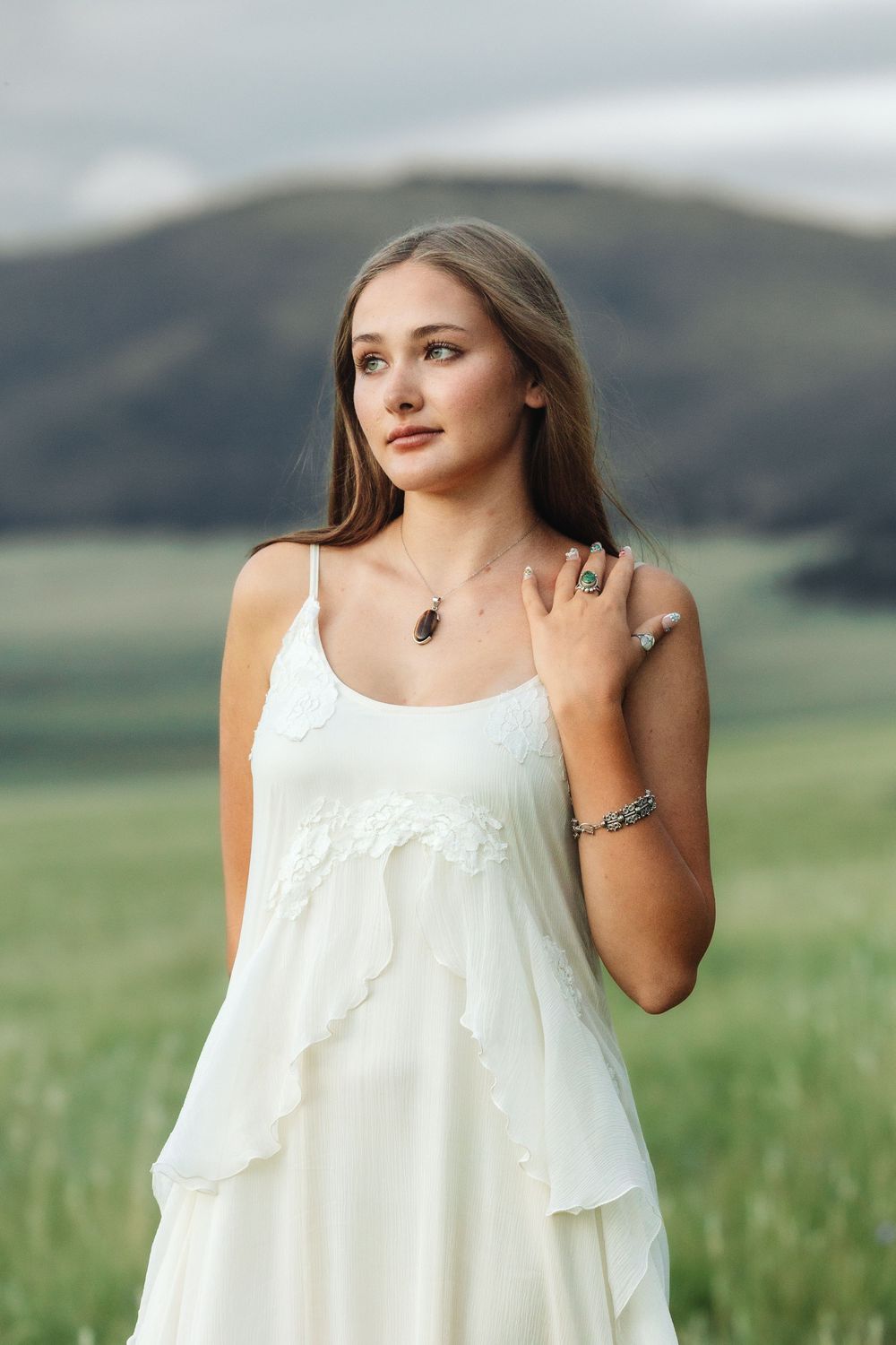 Young woman in white dress with jewelry poses in green field with mountains in background.