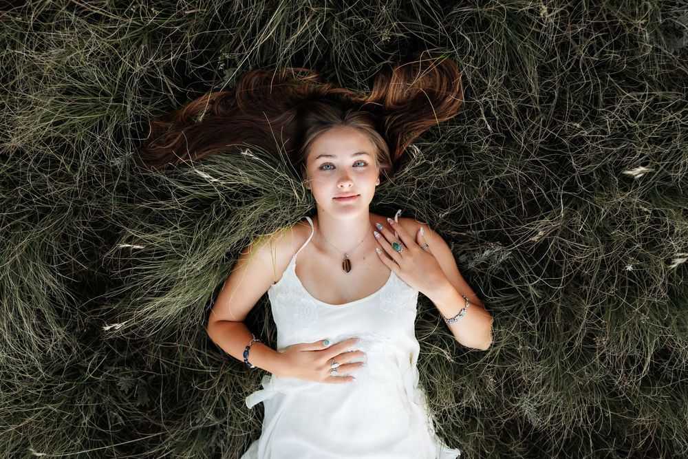 Young woman with long hair lying in green grass, wearing a white dress and delicate necklace, gazing upward peacefully.