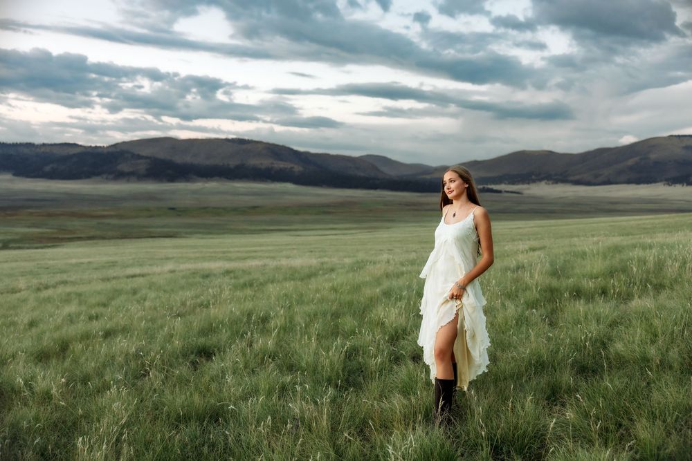 A woman in a white dress and boots stands in a vast green field with mountains and dramatic clouds behind her.