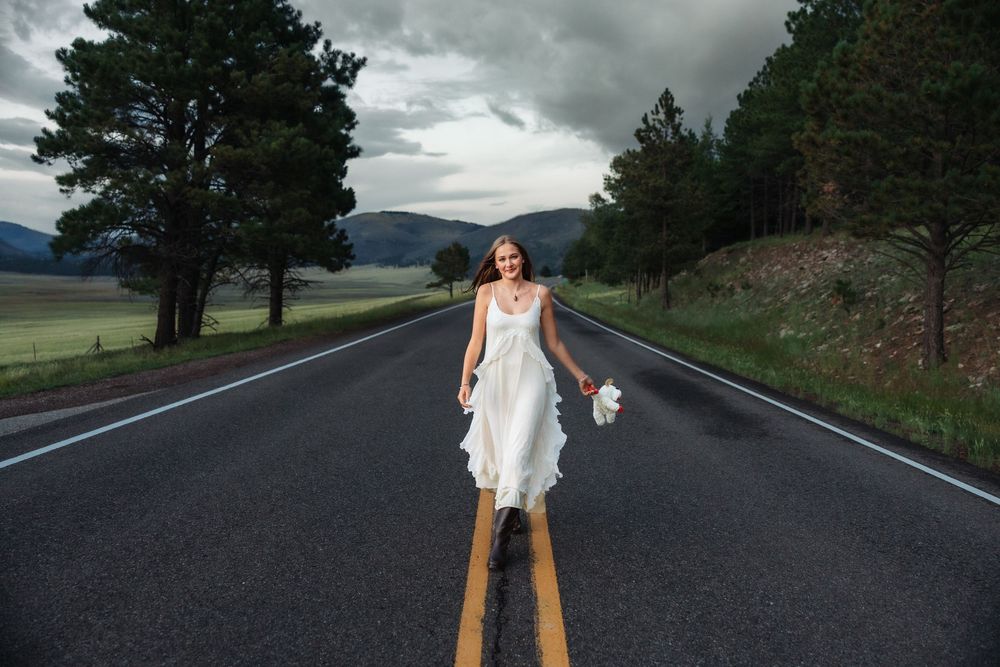 Bride in white dress walking on mountain road centerline holding bouquet under dramatic cloudy sky.
