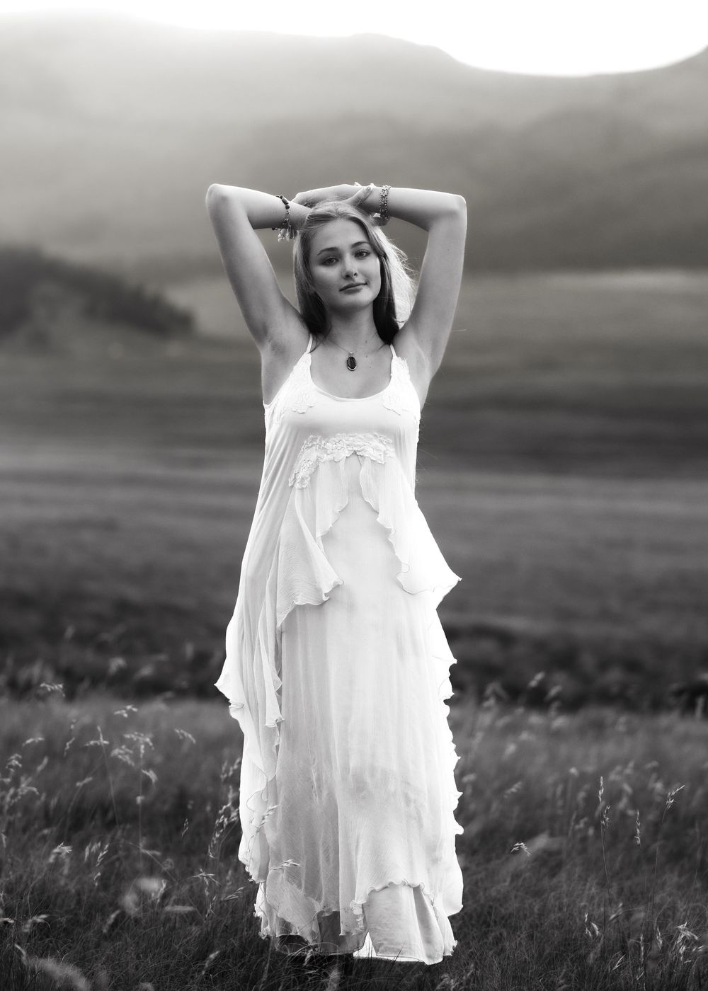Black and white photo of a young woman in a flowing white dress posing in a field with mountains behind her.