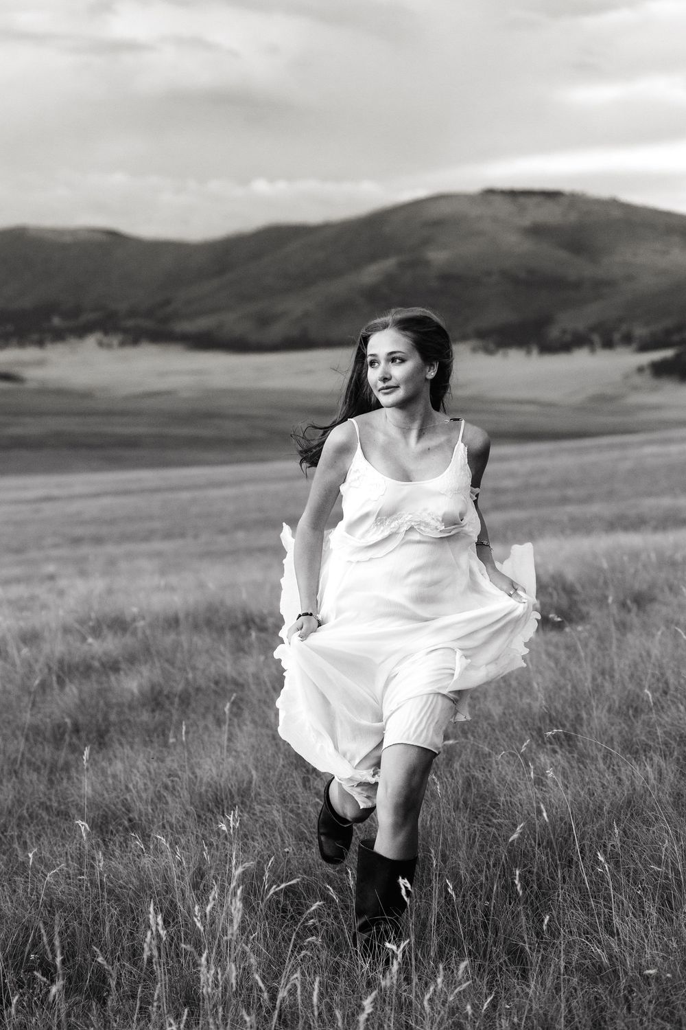 Black and white photo of a young woman in a white dress and boots running through a grassy field with mountains behind.