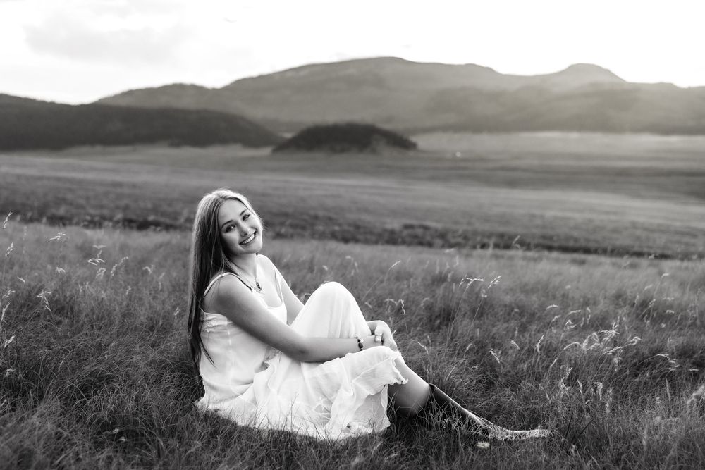 Young woman in white dress sitting in a grassy field, smiling, with mountains in the background, black and white photo.