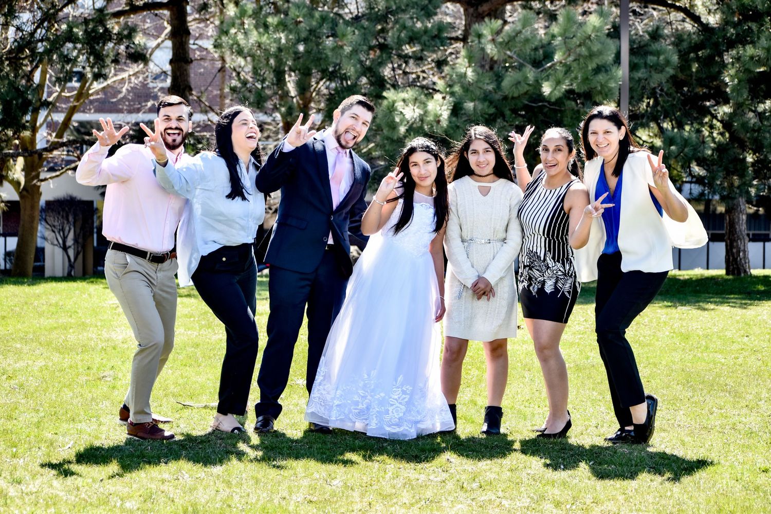 Wedding party posing on green lawn, bride in white gown with friends in formal attire, smiling outdoors.