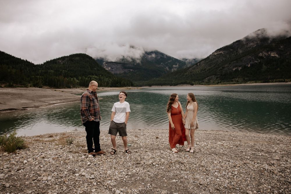 Family of four standing on rocky shore of a serene mountain lake with misty peaks in the background.