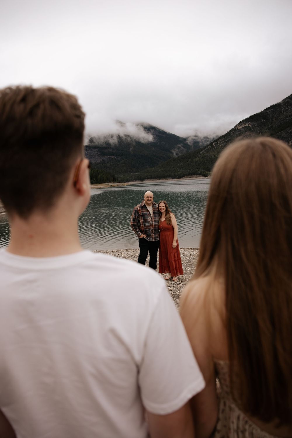 Couple posing by a mountain lake with misty peaks, viewed from behind two observers in the foreground.