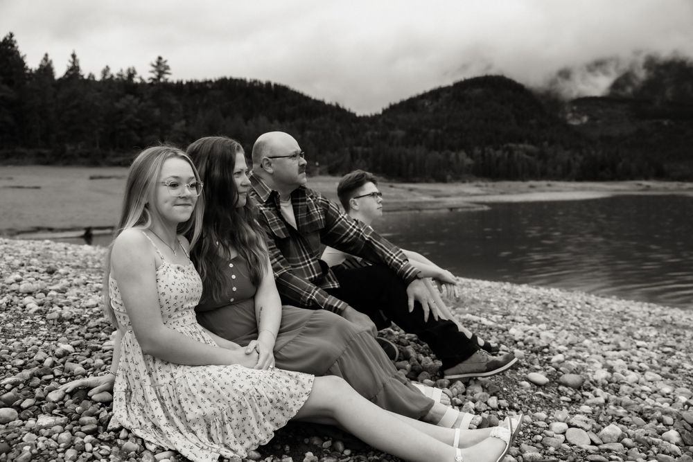 Black and white photo of four people sitting on rocky shore by a lake with misty mountains in background.