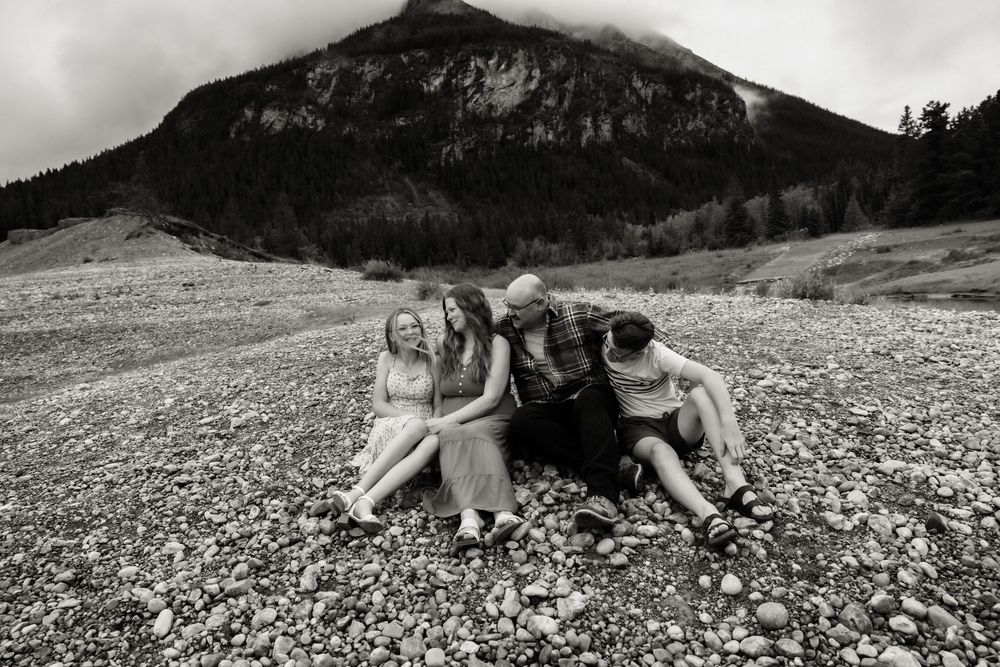 Black and white photo of a family sitting together on a rocky riverbed with a misty mountain in the background.
