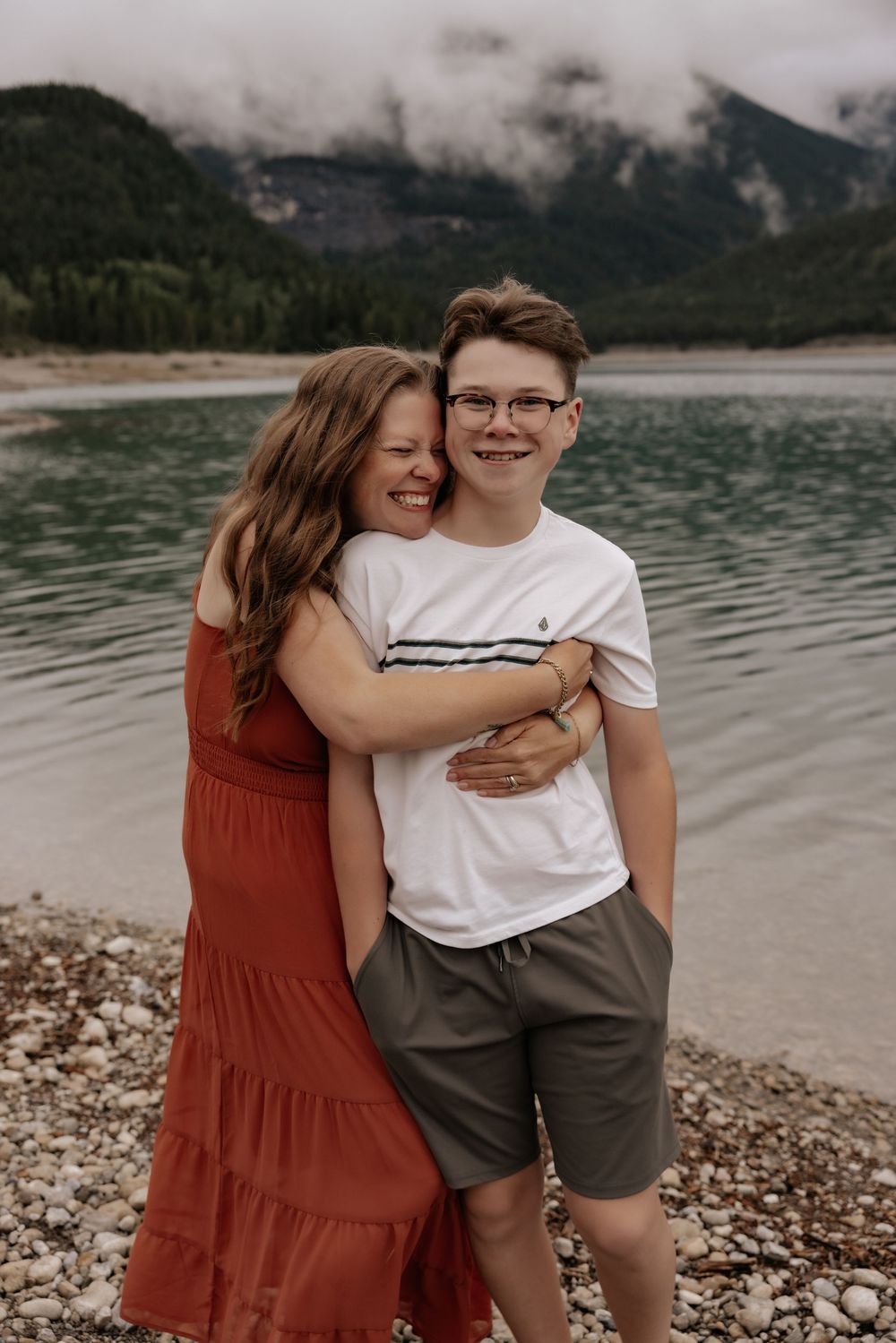 A woman in a red dress hugs a teenage boy in a white tee by a scenic mountain lake on a rocky shore.