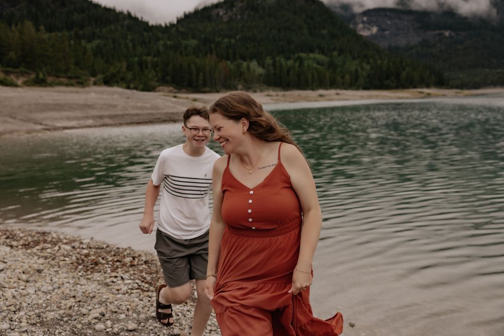Couple walking along a rocky lakeside shore with misty mountains in the background, woman in red dress smiling.