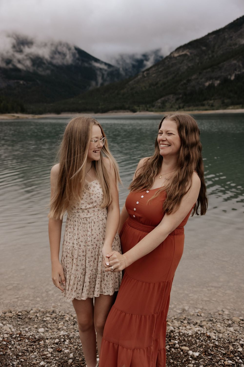 Two smiling women in summer dresses hold hands by a turquoise mountain lake with misty peaks in the background.
