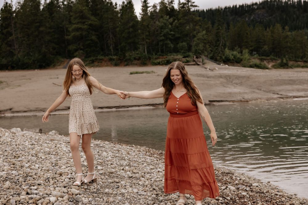 Two women in boho dresses hold hands and laugh on a rocky shoreline with evergreen trees behind them.