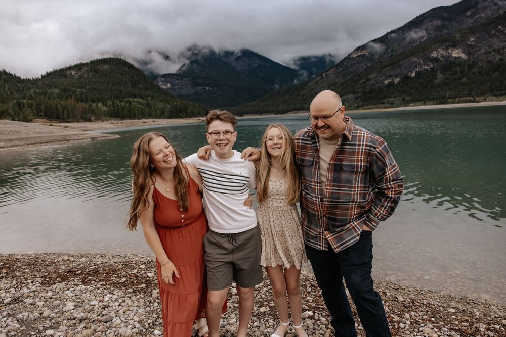 Family of four smiling together near a turquoise mountain lake with misty peaks in the background.