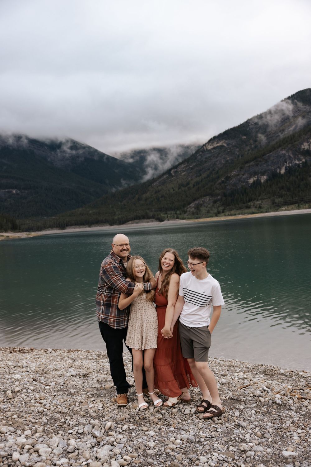 Family of four smiling together on rocky lakeshore in alberta Canada during thier family photoshoot