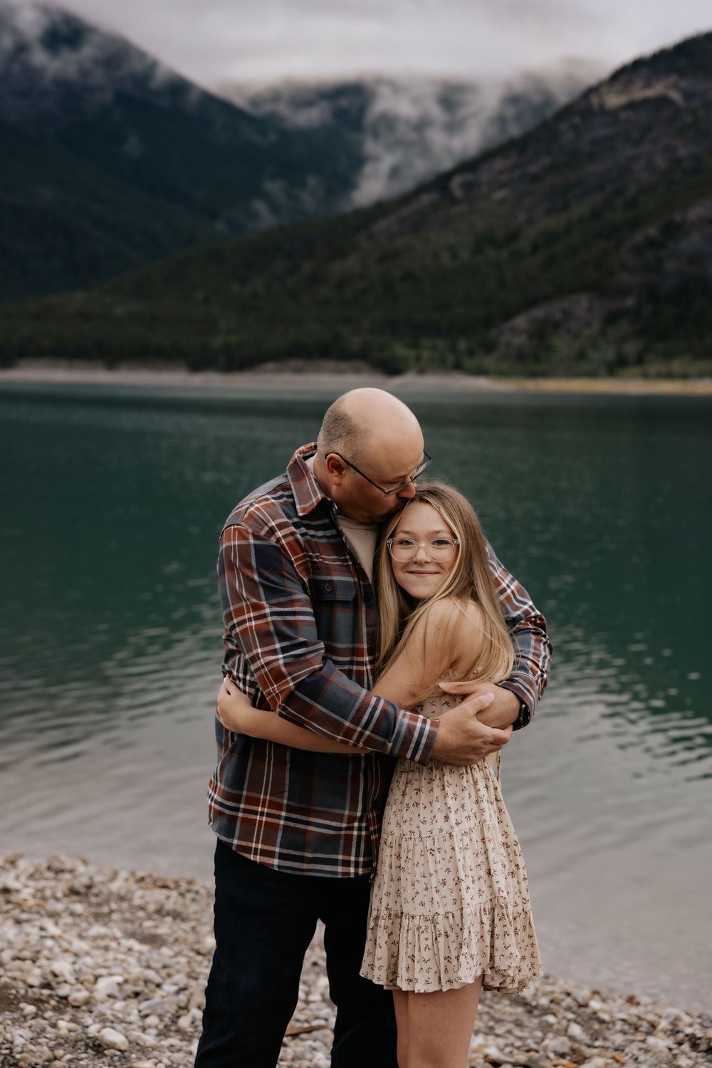 Couple embracing by a turquoise mountain lake, man kissing woman's cheek, rocky shore, misty peaks backdrop.