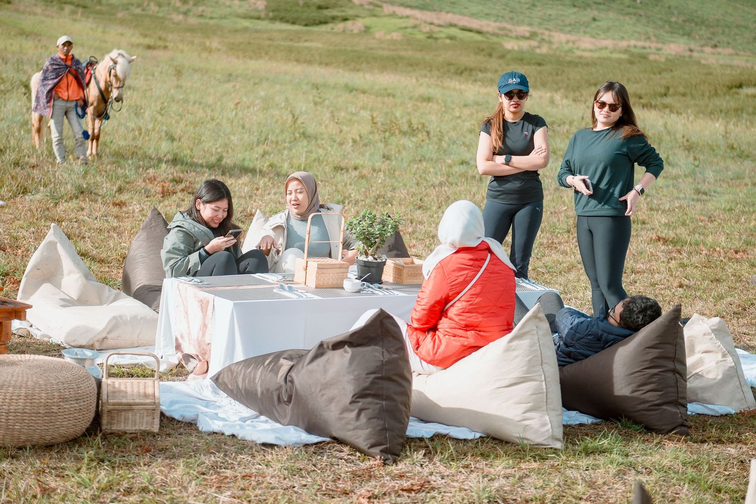 Group of women relaxing at outdoor picnic table with bean bag chairs in green meadow, horses visible in background.