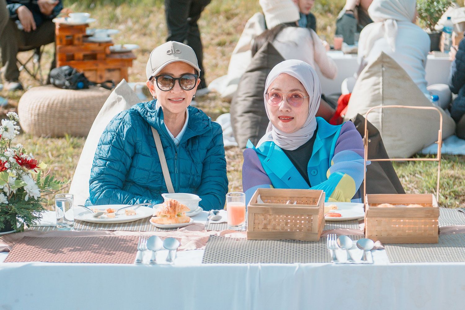 Two women smiling at an outdoor picnic table with wicker baskets, one in blue jacket and one wearing a white hijab.