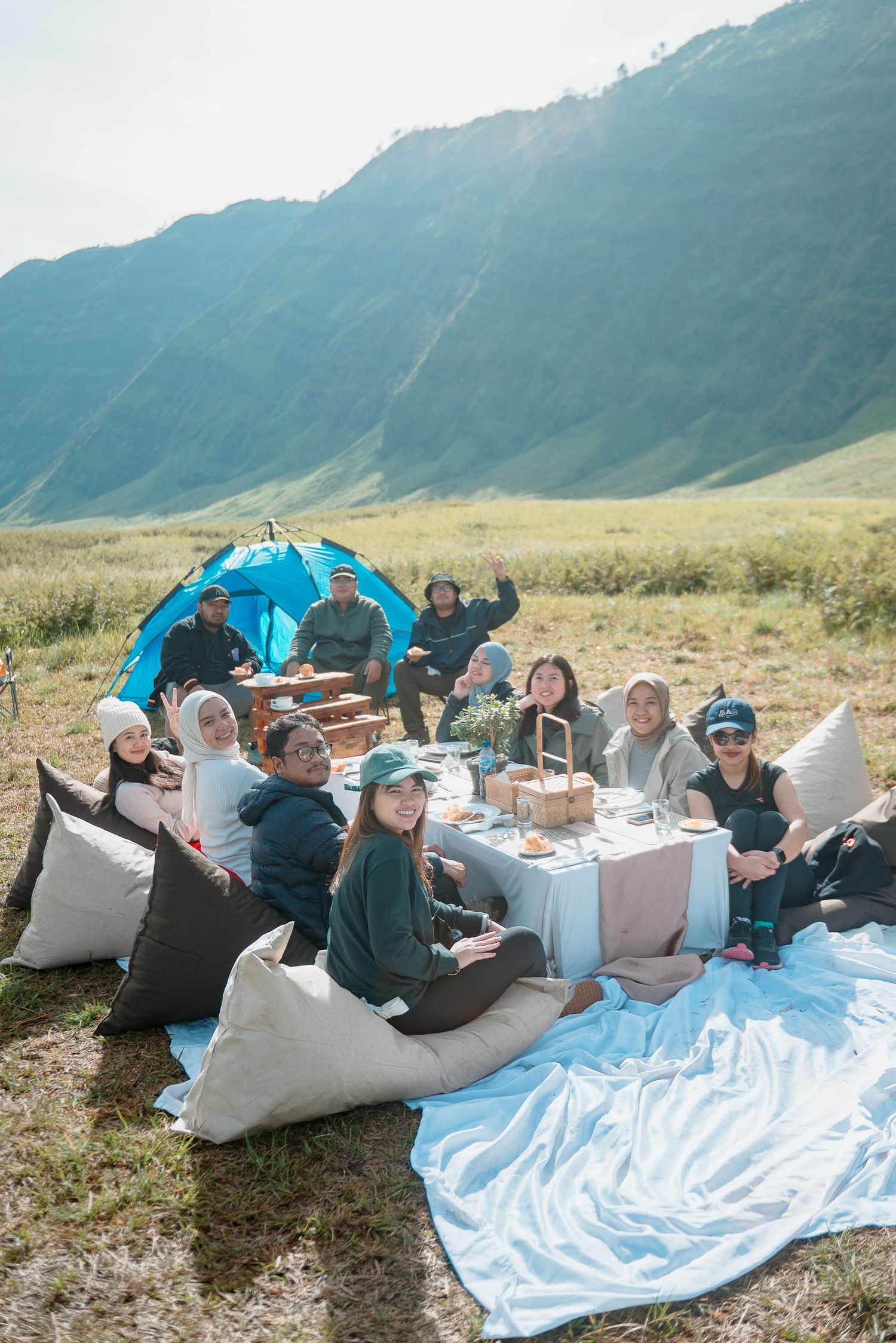 Group of friends enjoying a picnic on a mountain meadow with a blue tent and scenic hills in the background.