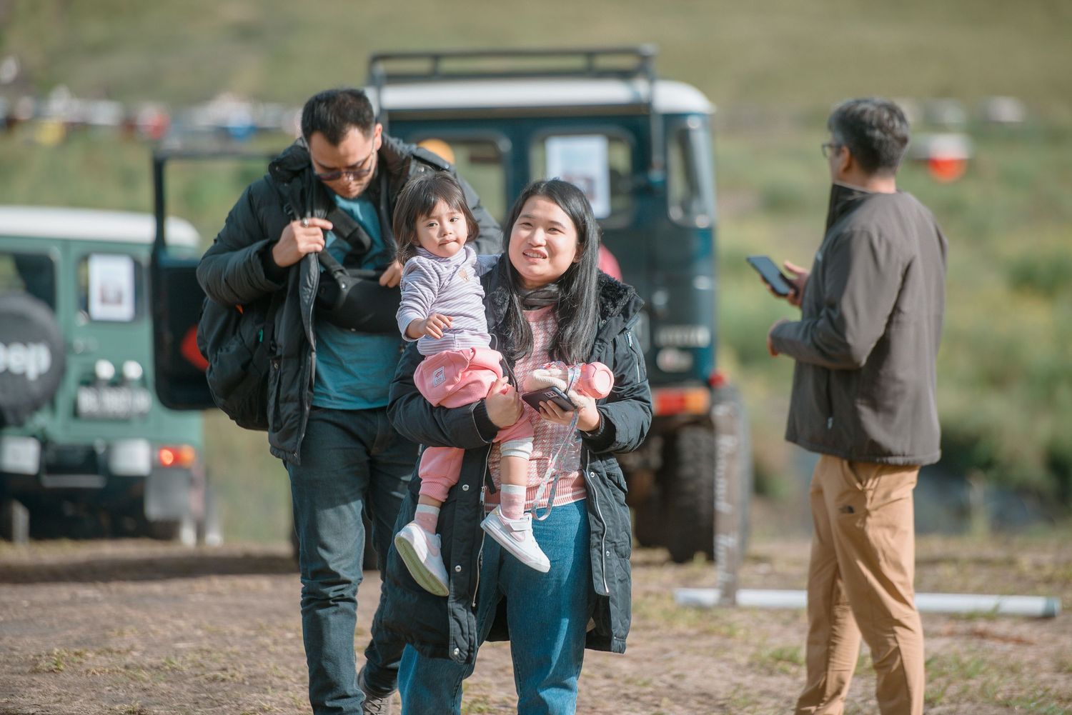 Family with young child smiling outdoors near a Jeep vehicle on a grassy terrain.