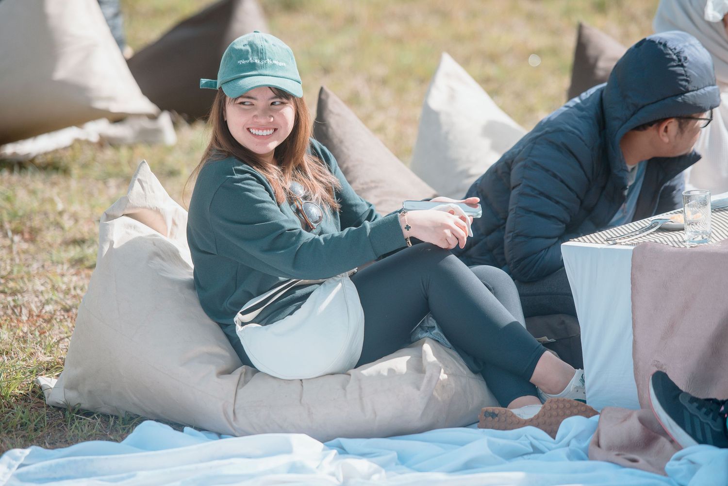 A smiling woman in a green cap relaxes on a beige bean bag chair outdoors.