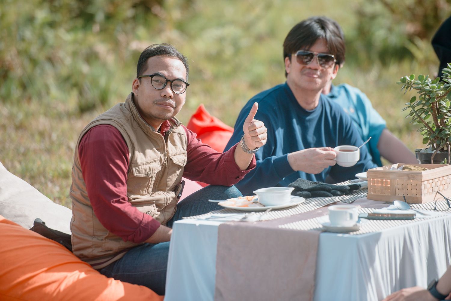 Two men sitting outdoors at a table with coffee cups, one giving thumbs up, enjoying a relaxed outdoor gathering.