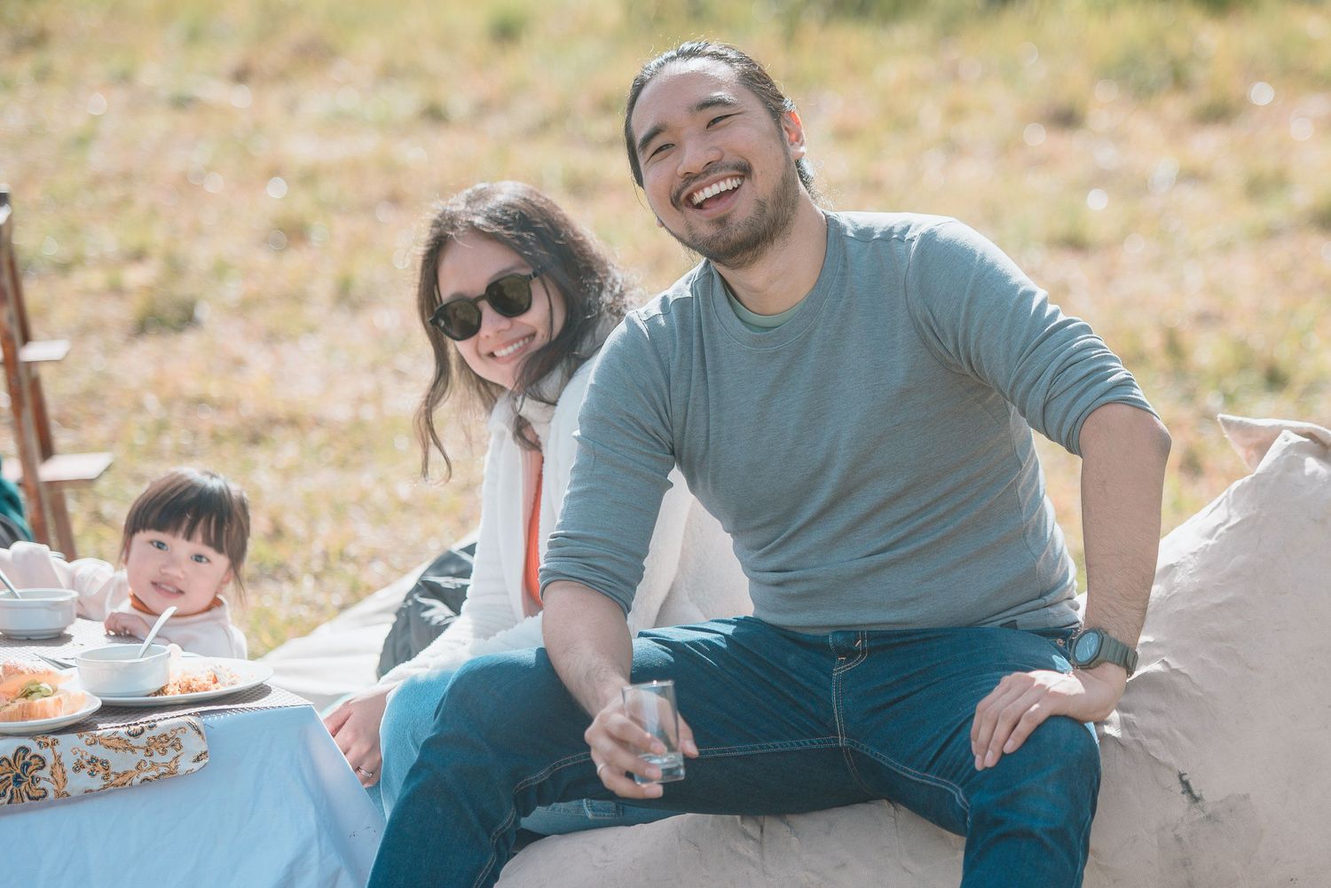 Family enjoying an outdoor picnic together, smiling couple with young child on a sunny day in a field.