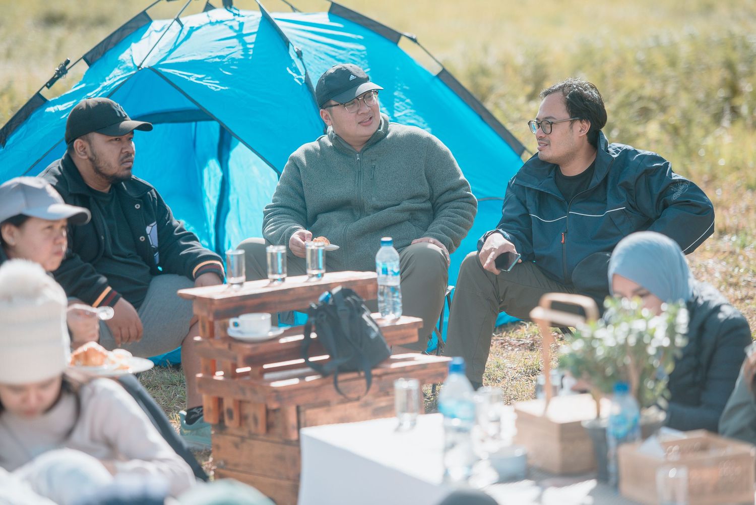 Group of friends camping outdoors, sitting near a blue tent, enjoying a picnic gathering in a scenic natural setting.