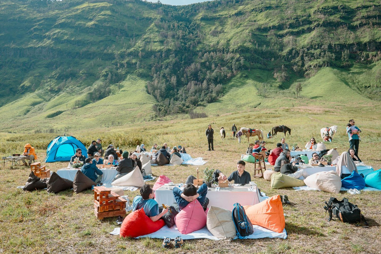Group of campers relaxing on colorful bean bags on a green mountain hillside with tents and horses in background.