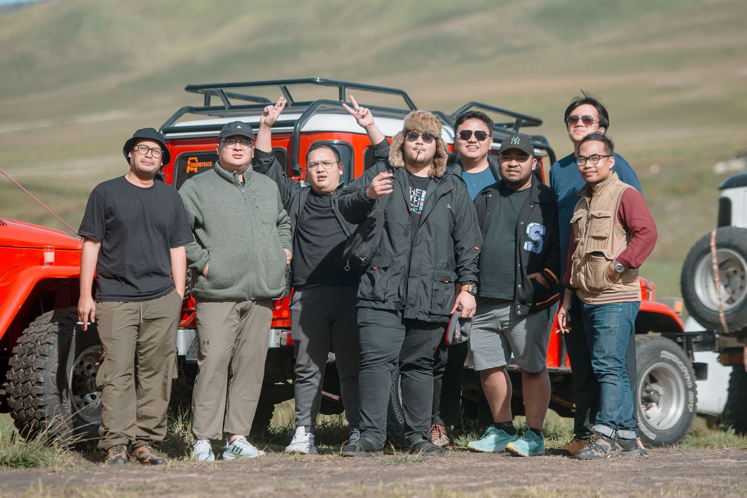 Group of people posing together outdoors near a red off-road vehicle in a scenic green valley landscape.