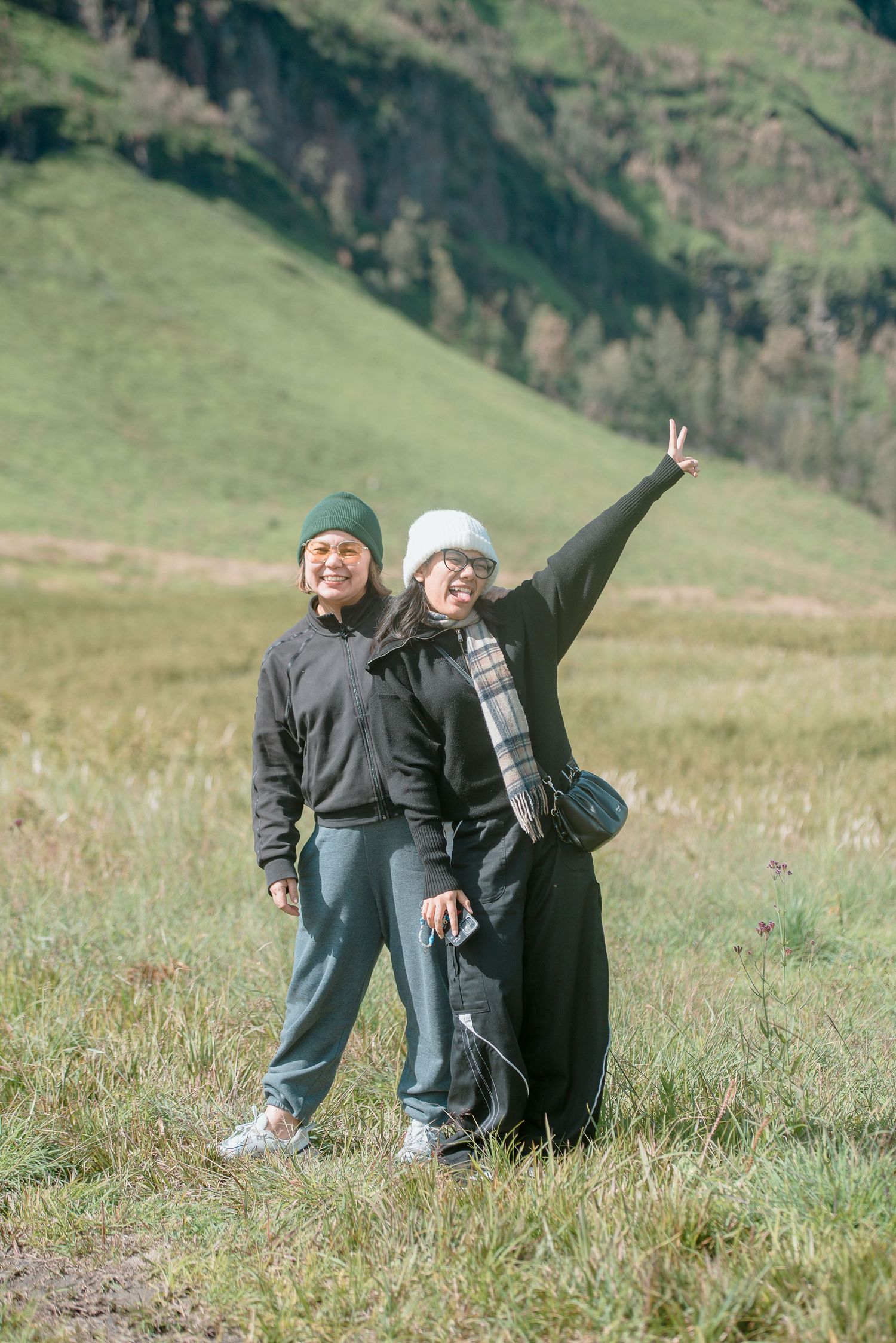 Two women in dark outdoor clothing and beanies pose happily in a green mountain meadow landscape.