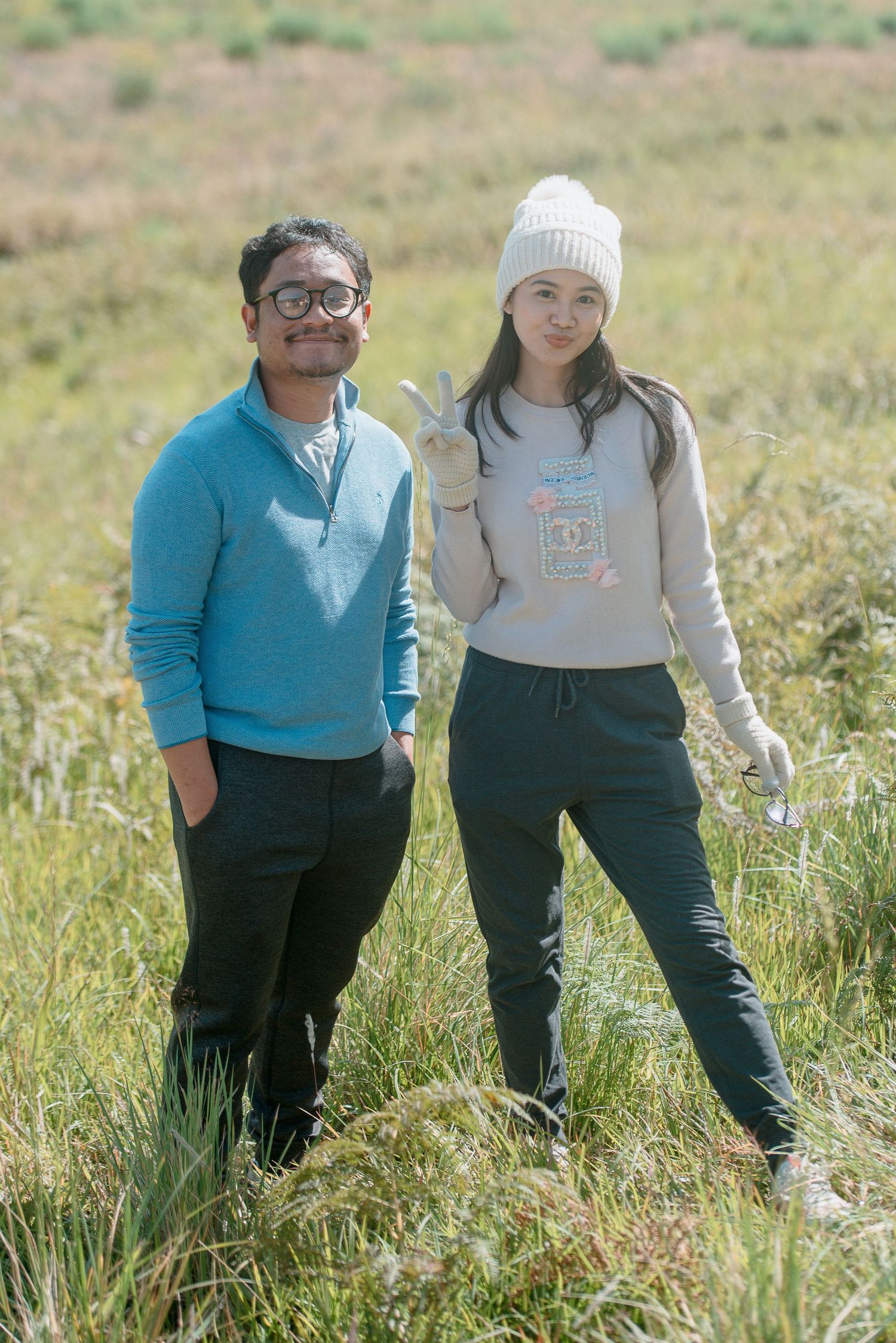 Two people standing in a grassy field, man in blue sweater and woman in beige hoodie and white beanie.