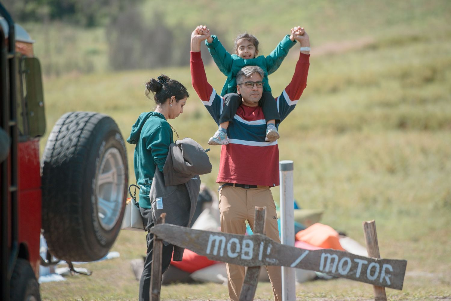 Family enjoying outdoor adventure near a Mobile Motor sign, child sitting on adult shoulders with arms raised.