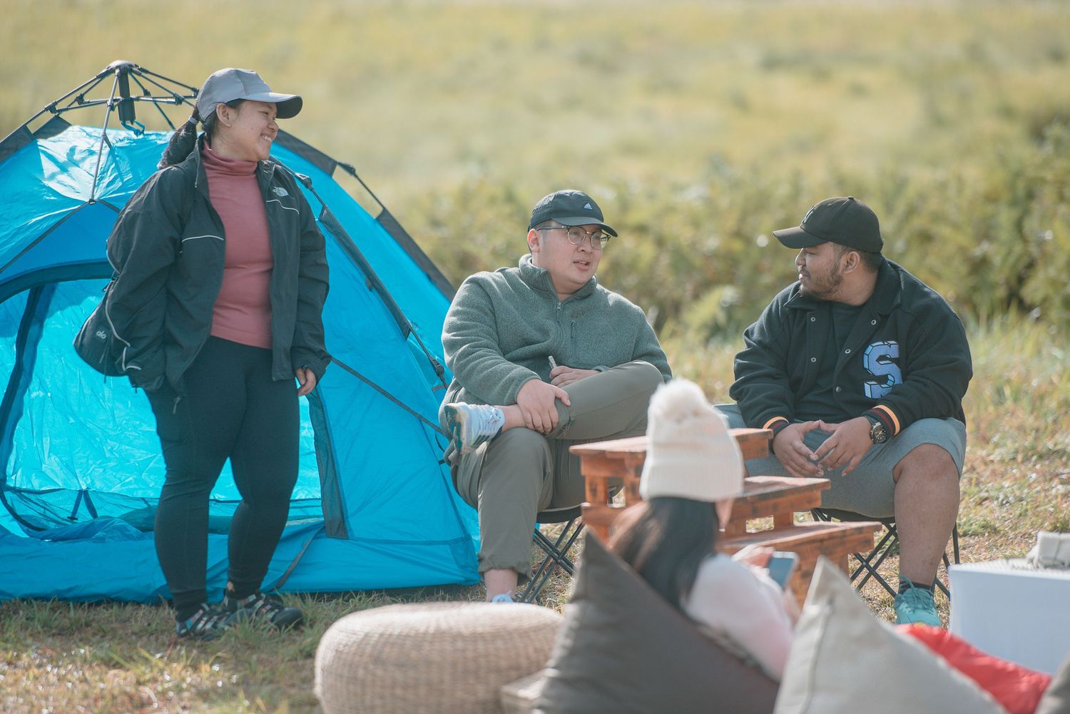 Group of friends camping outdoors, sitting near a blue tent in a grassy field, enjoying conversation.