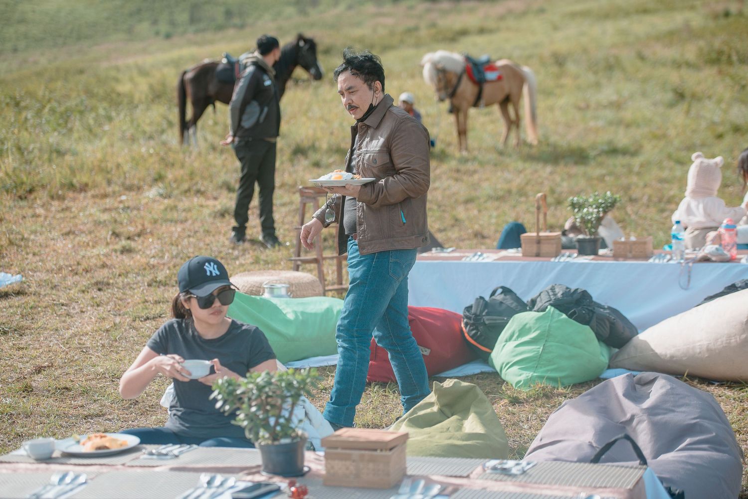 Two people enjoy an outdoor picnic on a blanket in a field with horses grazing in the background.