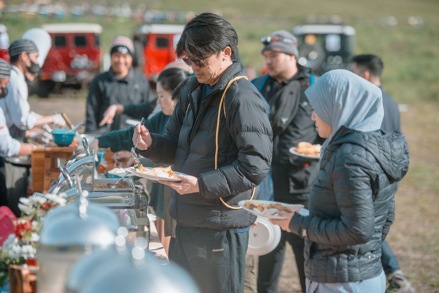 People serving themselves at an outdoor buffet event, wearing winter jackets, with vehicles visible in the background.