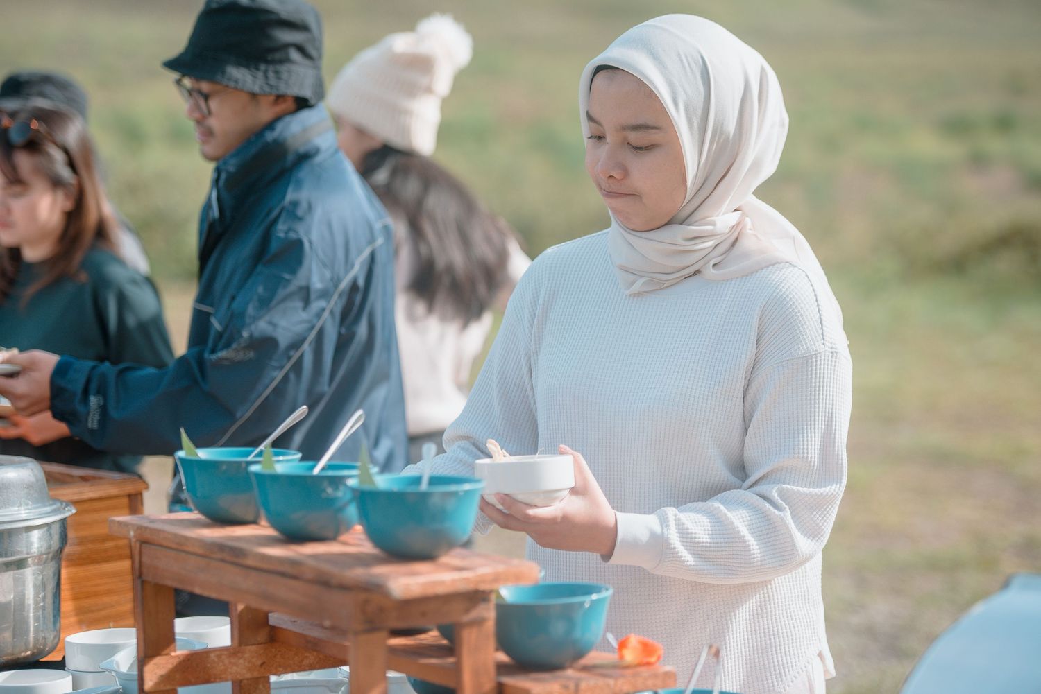 Woman in white hijab serving food outdoors at a buffet table with blue bowls in a scenic setting.