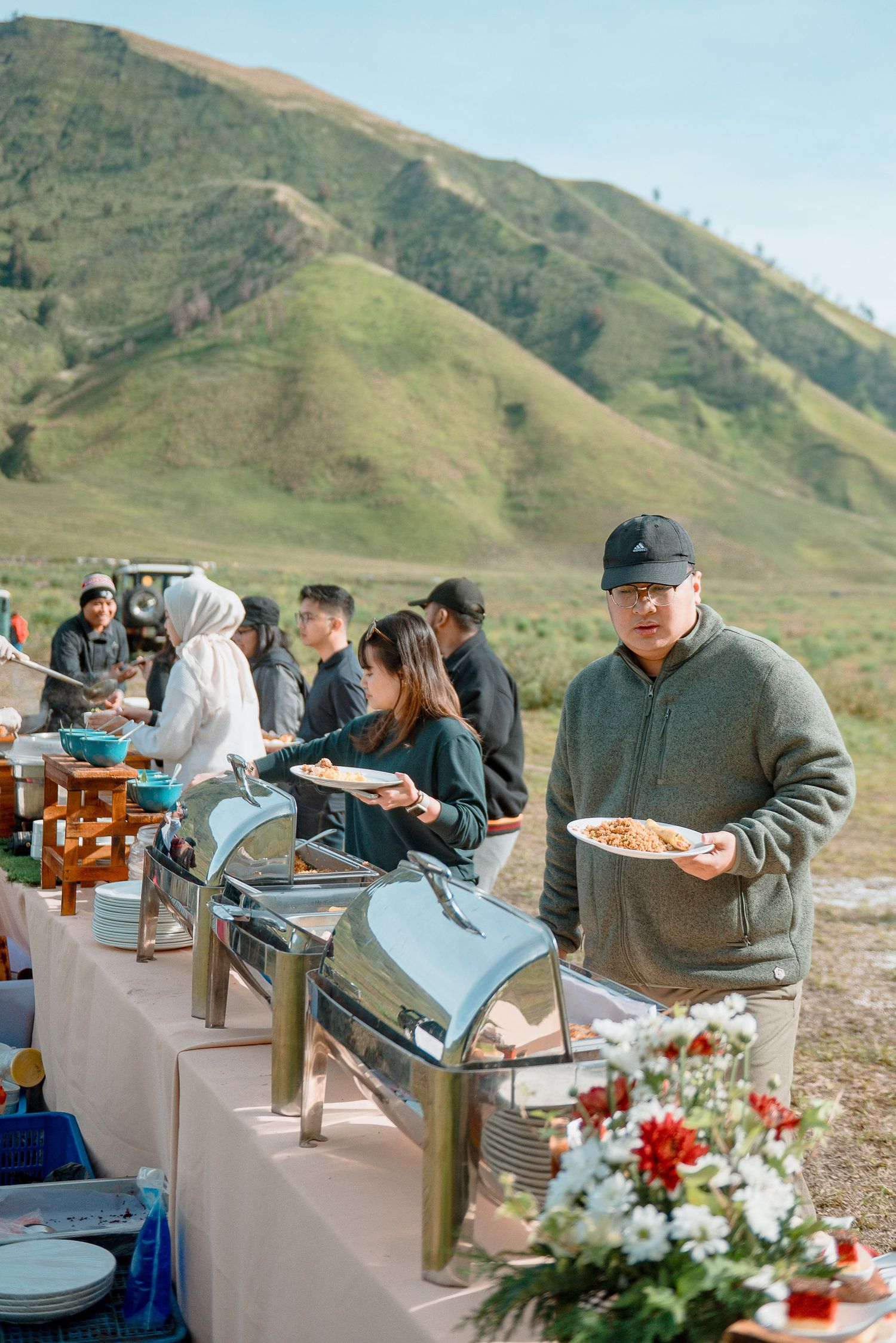 People serving themselves at an outdoor buffet with chafing dishes, set against lush green hills under a clear blue sky.