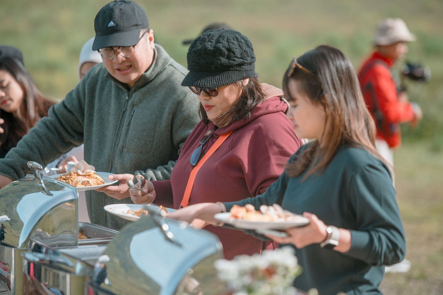 People serving themselves food at an outdoor buffet in a grassy field during a casual daytime gathering.