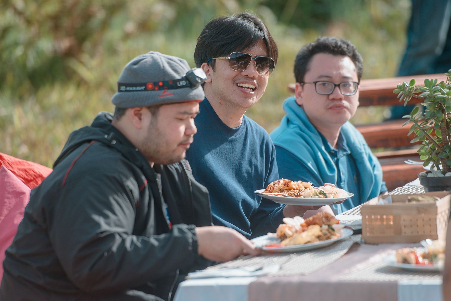 Three men sitting at an outdoor table enjoying a meal together, smiling and relaxed in a natural setting.