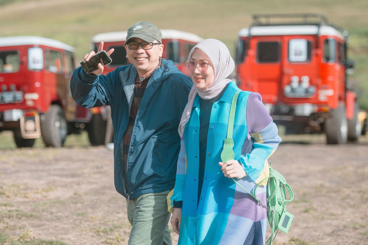 A couple smiles for a selfie outdoors with red off-road vehicles parked in the background.