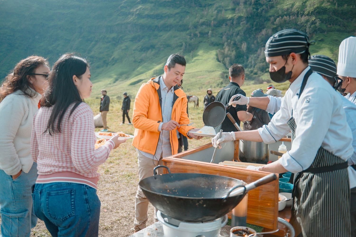 Chefs serving hot food from a wok to tourists at an outdoor cooking station in a scenic mountain valley.