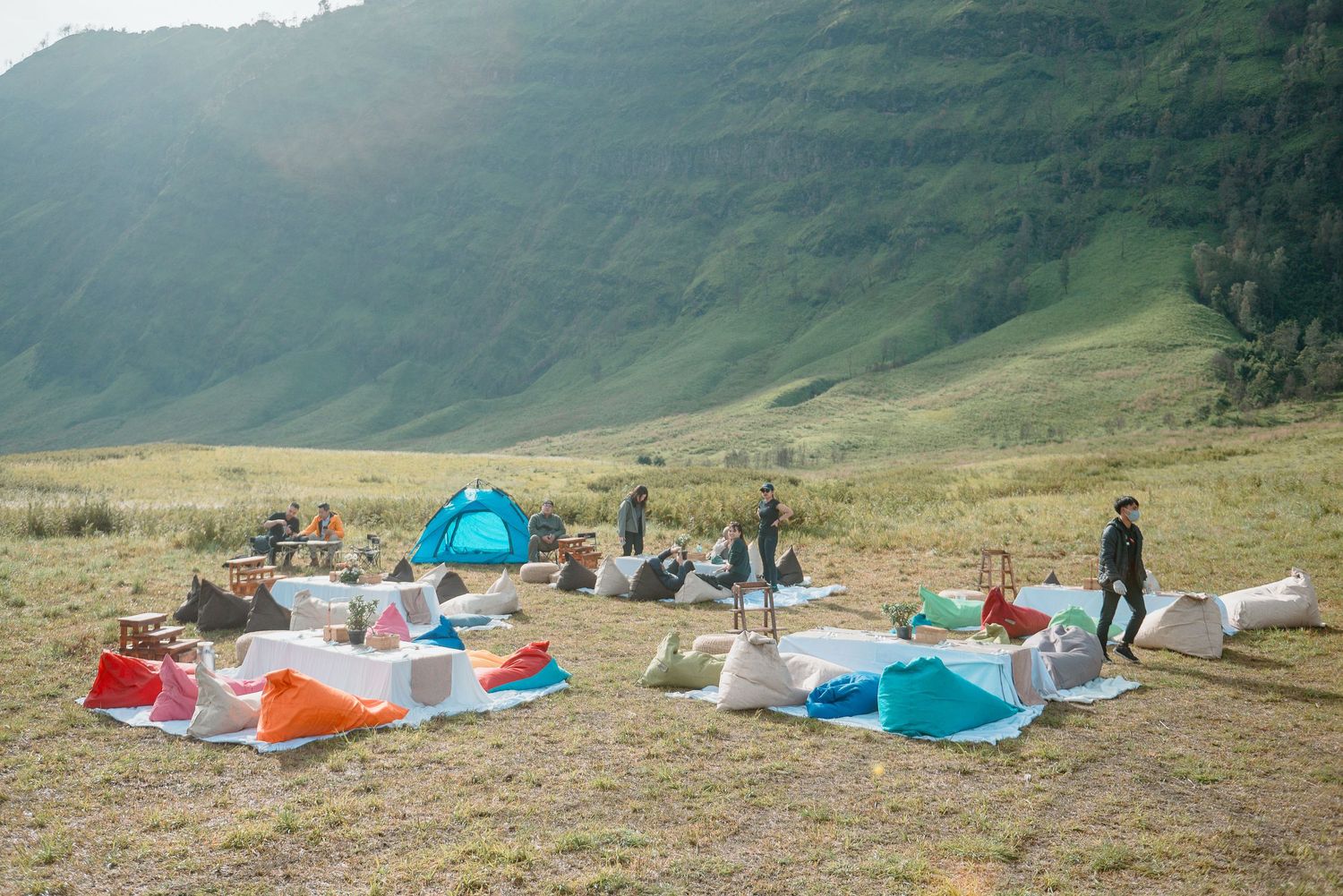 Colorful tents and camping gear spread across a grassy highland meadow with dramatic green mountain slopes in the background.