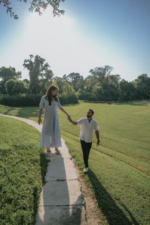 Couple holding hands walking along a garden path in a sunny park, woman in white dress, man in white shirt.